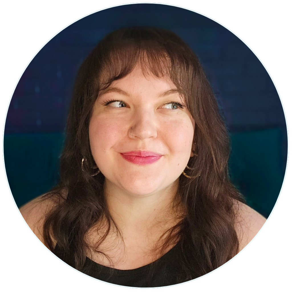 A picture of Laurel Holl, a white, fat woman with brown curly hair, heart shaped earrings, and on trend makeup smiling at the camera.